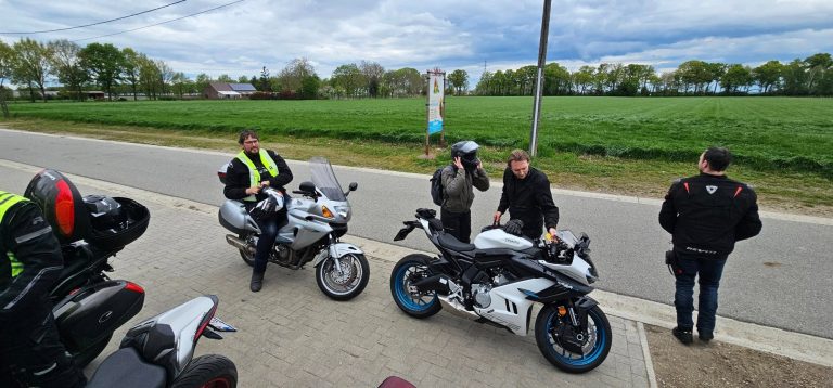 Twee motorrijders staan naast hun motoren langs de weg, met groen veld op de achtergrond.
